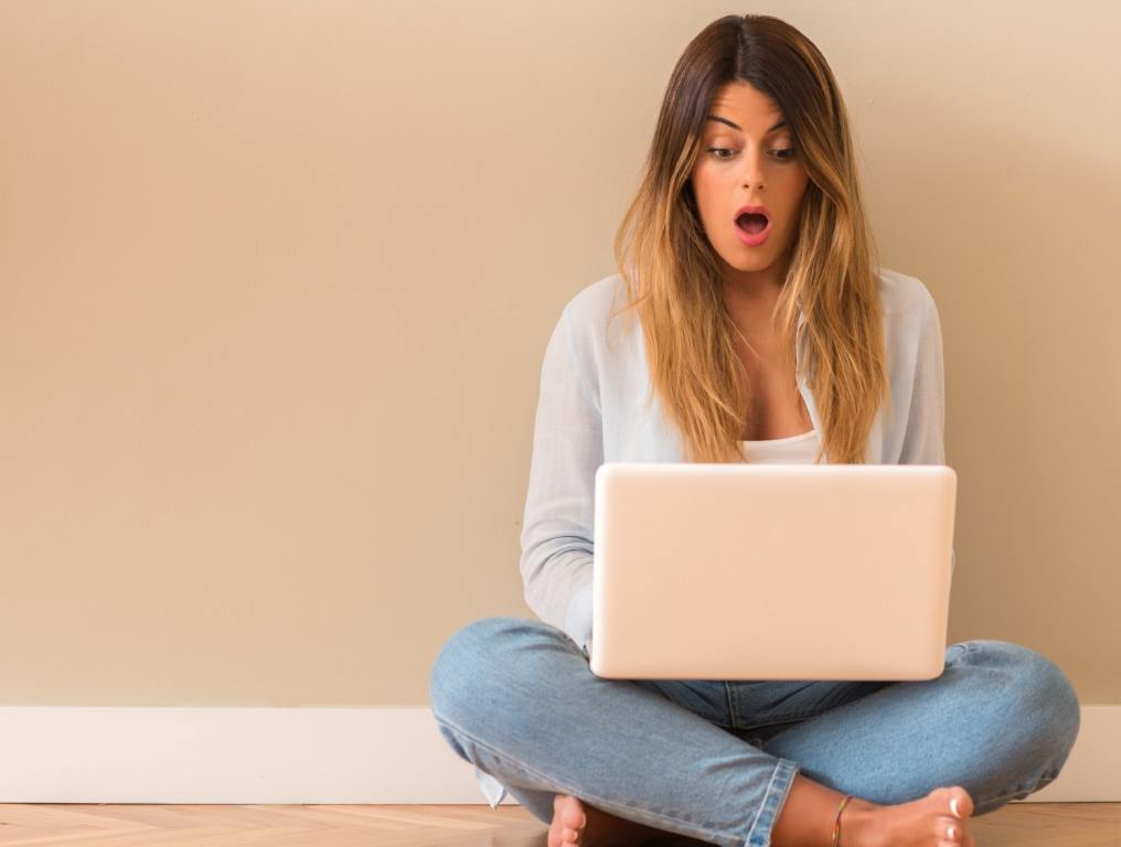 Young woman suprised sitting on the floor with computer looking at the laptop. (Red Flag Words To Avoid In Vacation Rental Listings)