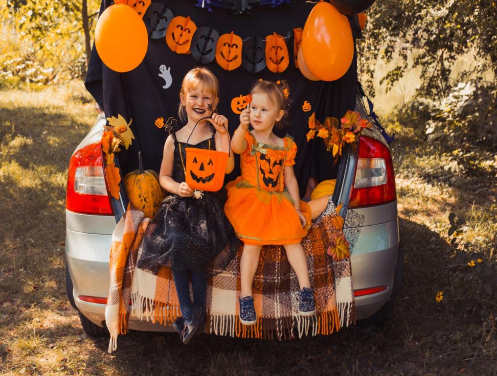 Two children in Halloween dresses sit on the trunk of a car that is decorated with Halloween decor. The older girl on the left is holding a pumpkin candy bucket and the smiling. The younger one on the right is holding up a sucker. Vegas Halloween.