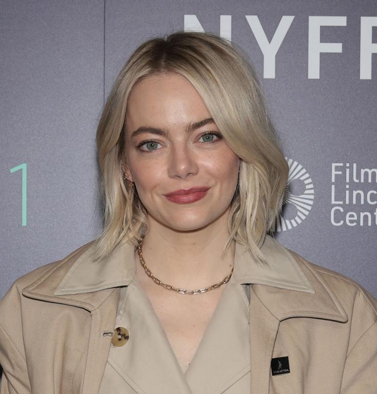 Emma Stone attends "Bleat" during the 61st New York Film Festival wearing a khaki trenchcoat smirking with platinum blonde hair.