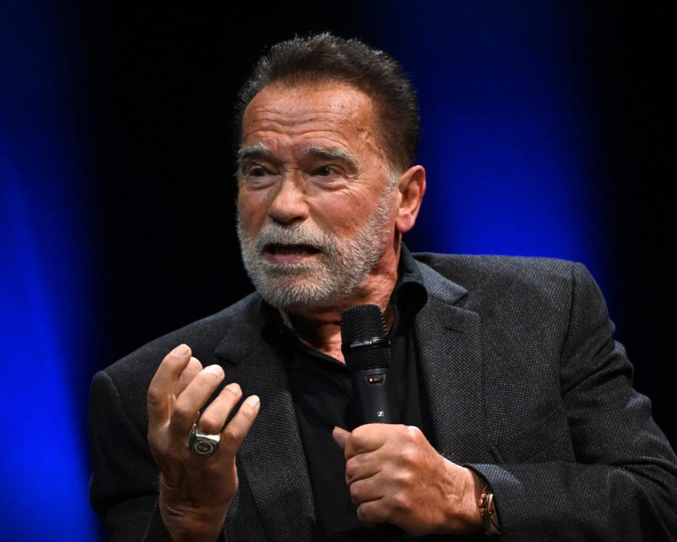 Arnold Schwarzenegger speaks onstage at an Evening with Arnold Schwarzenegger wearing a blazer facing left with one hand holding a microphone.