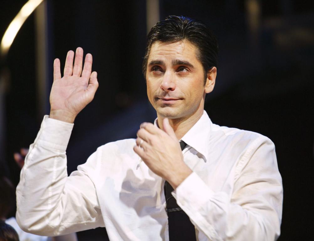 John Stamos joins the cast of "Nine" raising one hand up while the other caresses his chin on stage wearing a white button up and black tie.