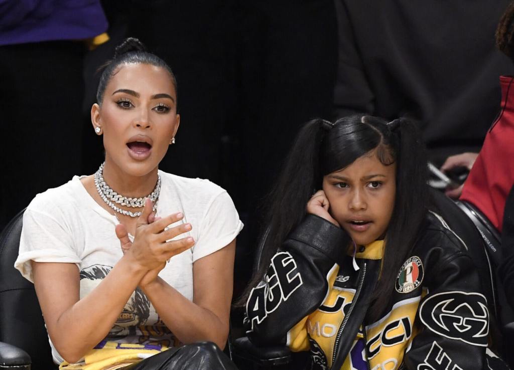 Kim Kardashian clapping wearing a white graphic tee with her mouth open and daughter North West with her hand on her cheek wearing a Gucci jacket attends the Western Conference Semifinal Playoff game