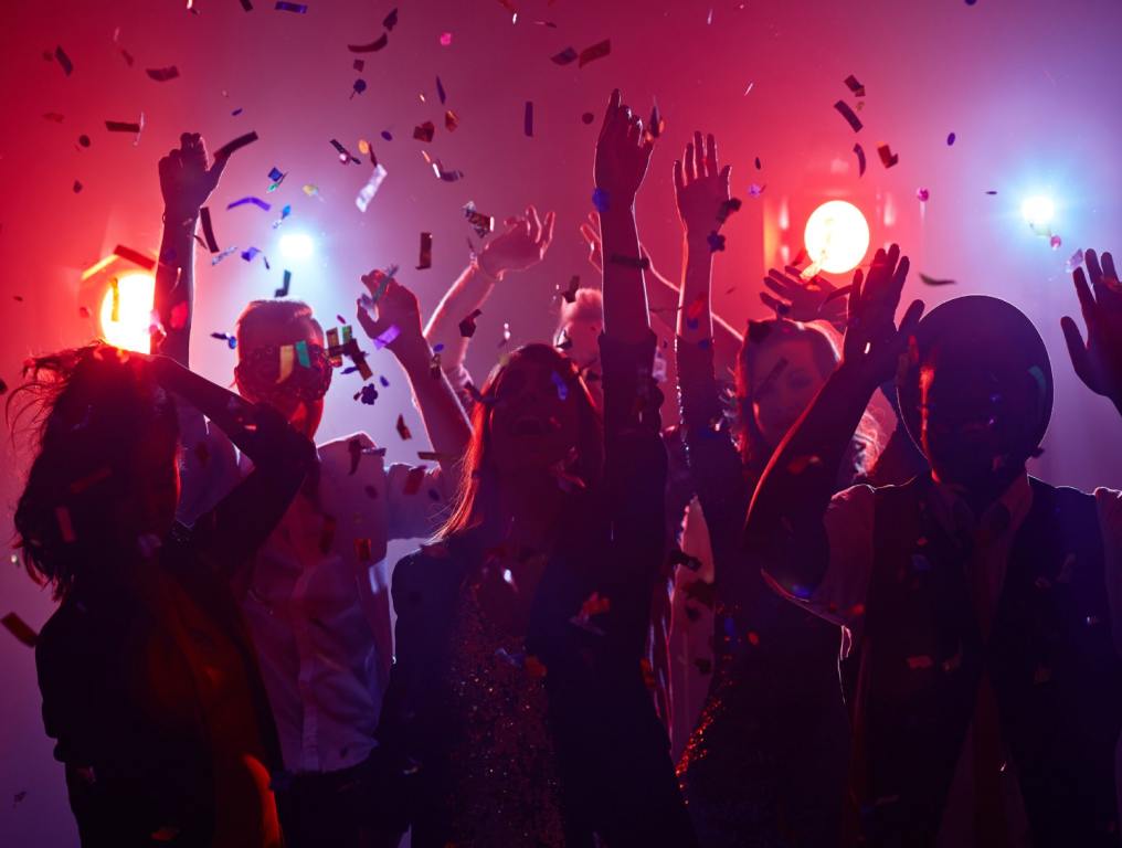 Friends are throwing their hands up at a New Year's Eve concert in Las Vegas. Confetti is falling from the sky and the people are seen from the back with stage lighting behind them.