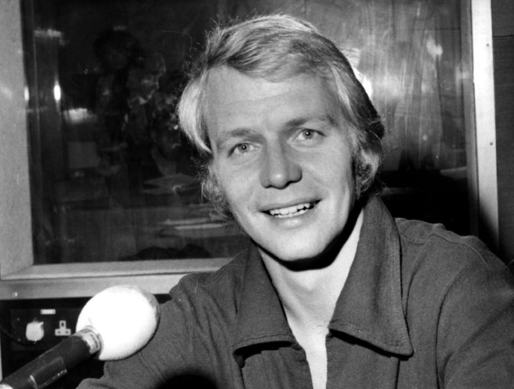 Actor David Soul in a black and white photo from the 1970s smiling in front of a mic with a dark shirt on