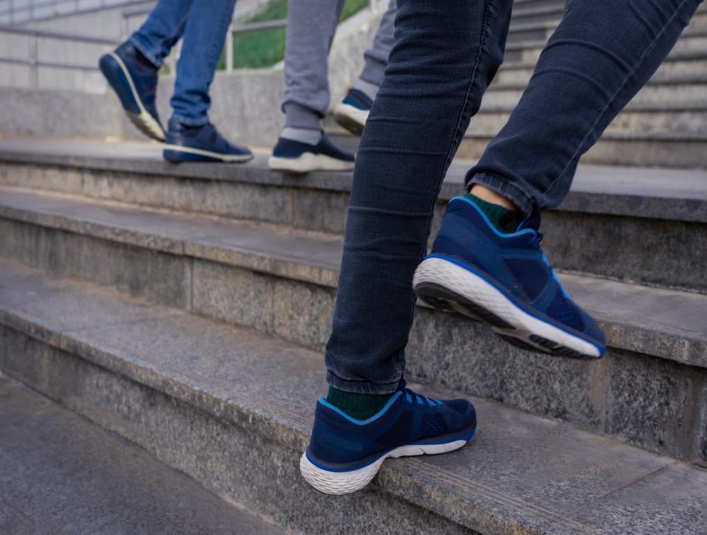 Cropped photo of the feet of three people climbing stairs. They are wearing jeans and tennis shoes. The stairs are outdoors and made of gray concrete.