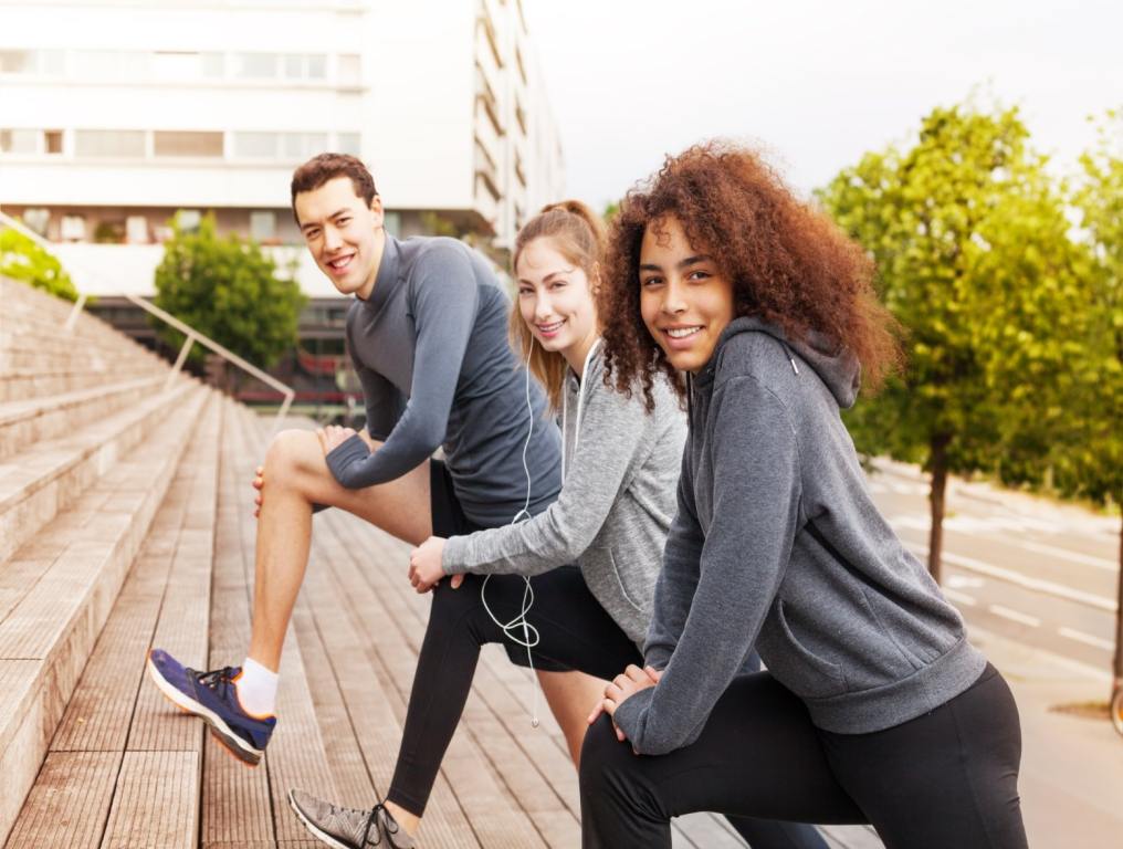 Two young woman and a young man stretch on a set of outdoor stairs. They are dressed in fall workout clothes and smiling at the camera. Stairs Las Vegas.