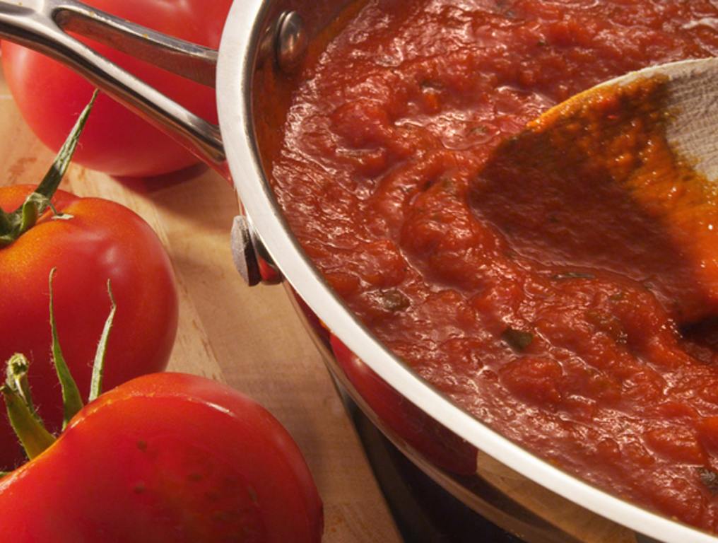 Marinara sauce simmering in a pot. Wood spoon seen stirring the sauce. There are fresh uncut tomatoes on the table beside the pot of marinara sauce.