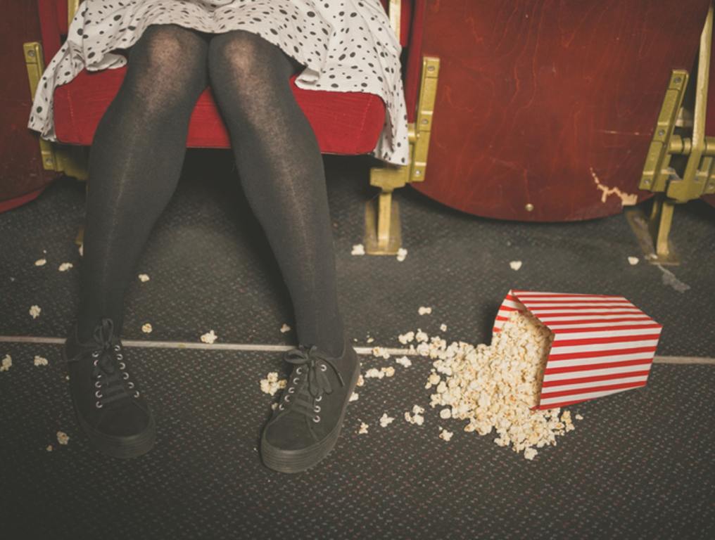 A young woman is sitting in a movie theater with a bucket of popcorn spilled on the floor in front of her