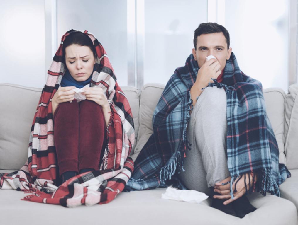 The couple is sitting on the couch wrapped in blankets. Man and woman are sick. The man flies out. Woman holding napkin, being sick, getting sick concept.