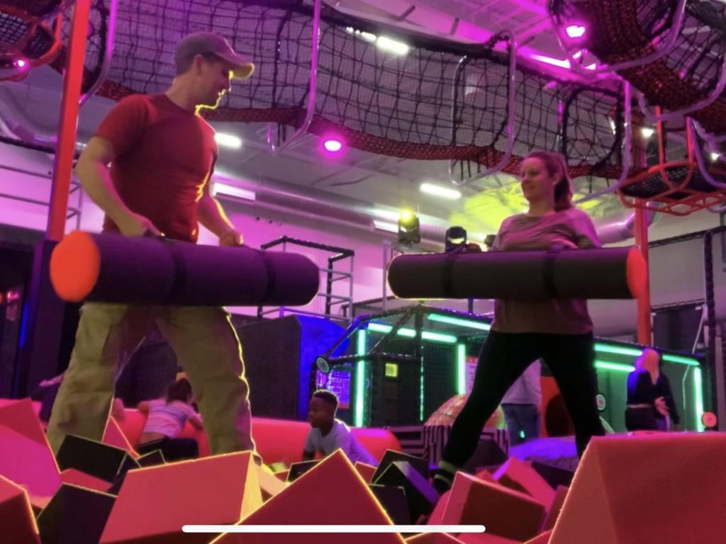 A man and woman stand on platforms in a pit full of foam cubes. They are each holding giant jousting cylinders. Staring at each other, reading to engage in combat.