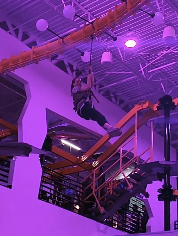 A woman in black leggings and brown t-shirt is harnessed to a red zipline and is suspended in mid air.