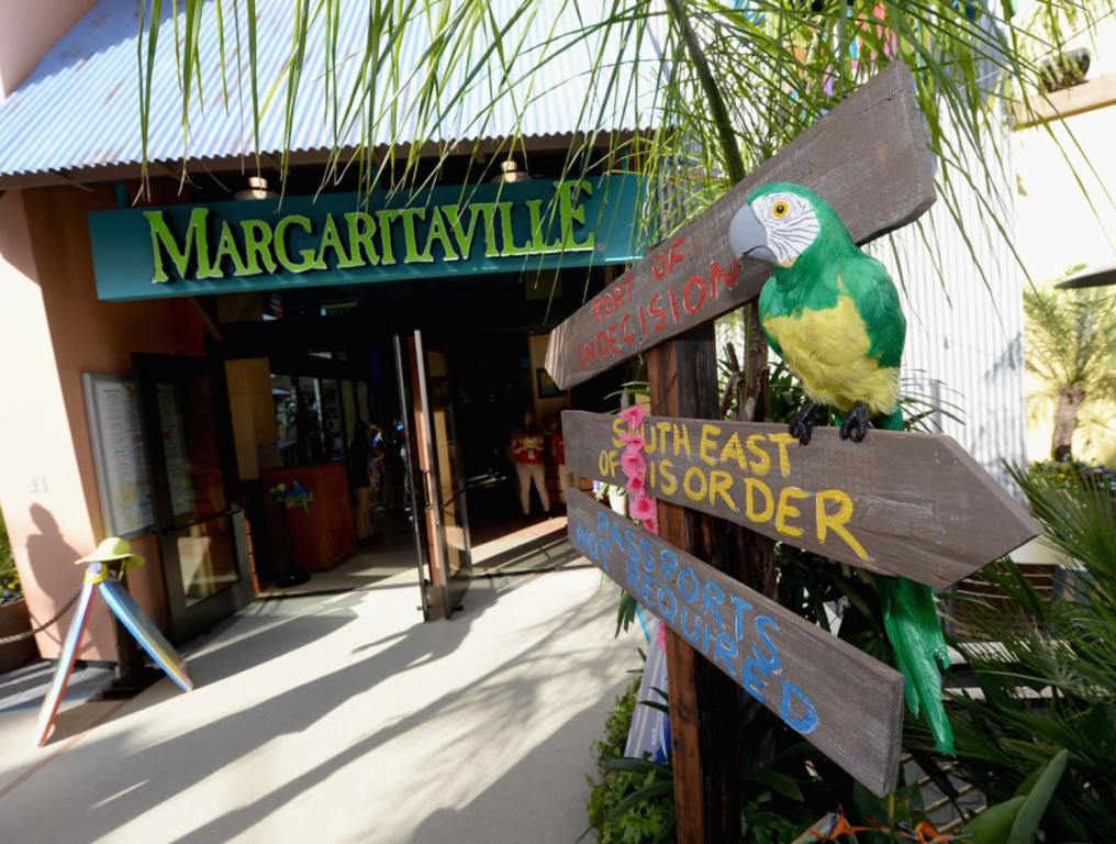 Photo of the entrance to Jimmy Buffett's Margaritaville restaurant at Universal Studios Hollywood. The double door entrance doors are wide open. Above the doors, the Margaritaville sign can be seen in bright green letters on a dark green background. Prior to the doors on the right hand side of the photo there are signs which read "Passports Not Required" on the bottom, "South East of Is Order" in the middle, and "Port of Indecision" on the top. Perched on the right edge of the middle wooden sign is a colorfully painted wooden parrot.