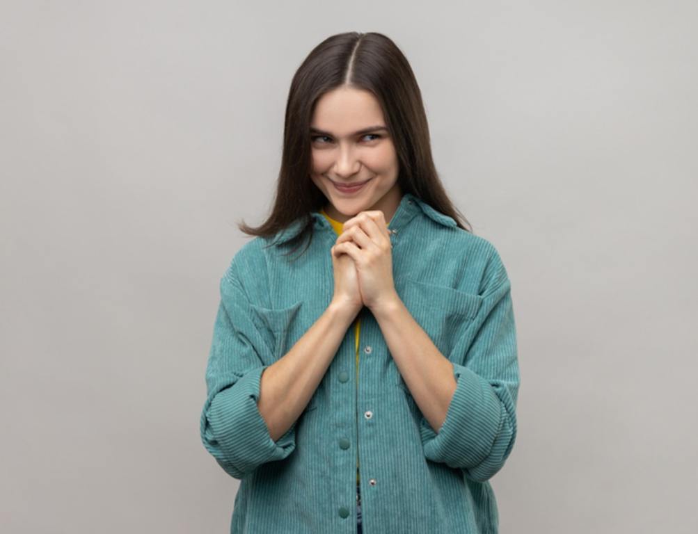 Devious cunning young woman with dark hair clasping hands and smirking mysteriously, scheming cheats, evil prank, wearing casual style jacket. Indoor studio shot isolated on gray background.