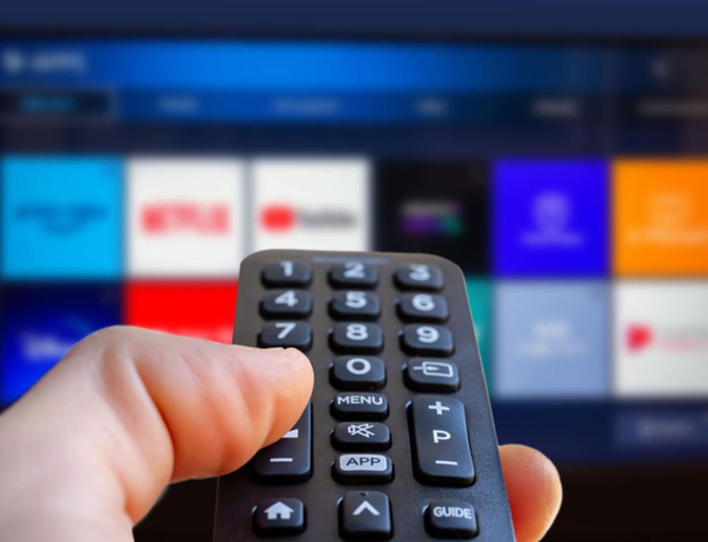 A man is holding a remote control of a smart TV in his hand. In the background you can see the television screen with streaming entertainment apps for video on demand, X