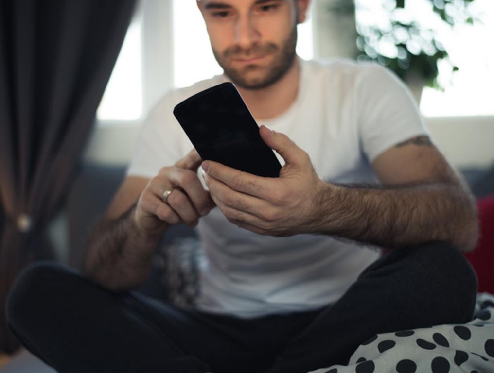 Young cell phone addict man in bed using smartphone, incognito mode concept.