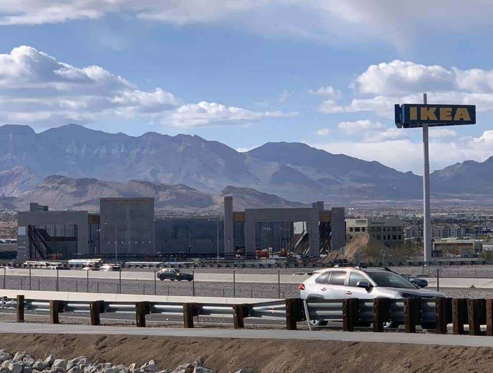 The Las Vegas IKEA sign stands next to a concrete structure under development. Cars passing on a freeway are shown in the foreground and mountains and clouds in the background.