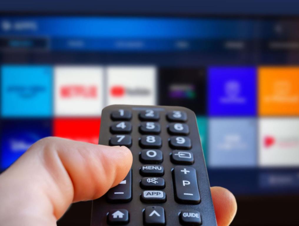 A man is holding a remote control of a smart TV in his hand. In the background you can see the television screen with streaming entertainment apps like disney+ for video on demand