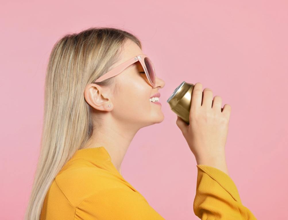 Beautiful happy woman drinking from beverage can on pink background