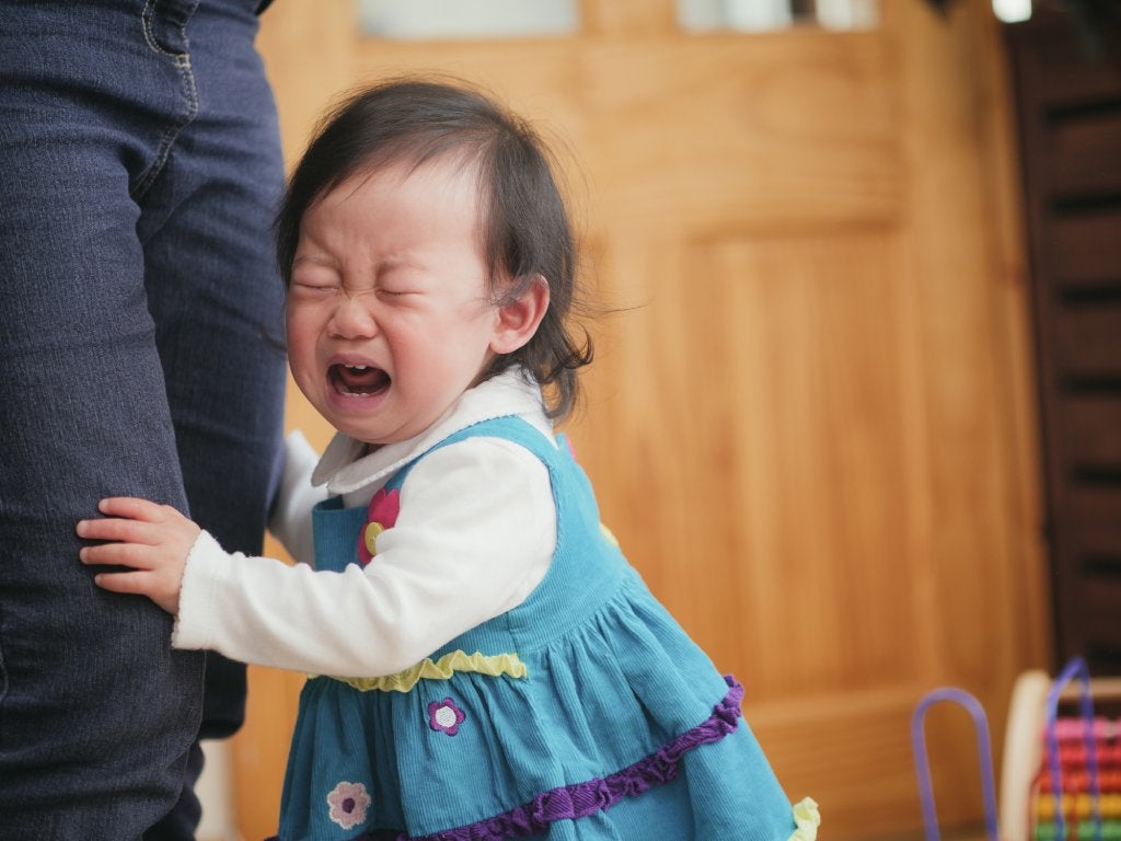 An Asian toddler girl in a blue dress and white shirt is clasping onto her parent's leg. They are standing in a kitchen and the parent is wearing blue pants.