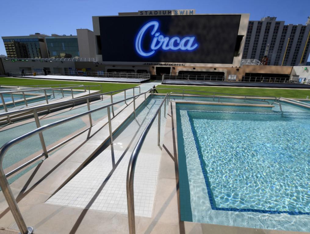 A wide-angle view of the Stadium Swim rooftop pool, the site of this year's Ultimate Graduation Do-Over Party for Class of 2020 CCSD high school graduates. Photo taken on a sunny day in downtown Las Vegas before the hotel's grand opening in October 2020. The "Circa" logo is featured in blue script on the 40-foot tall high-definition video screen at the far end of the complex. Above the screen is a concrete sign with dark blue letters reading "Stadium" and light blue letters reading "Swim." Two of the six pools can be seen in the foreground of the photo. There cement walkways with chrome hand rails between the two pools.