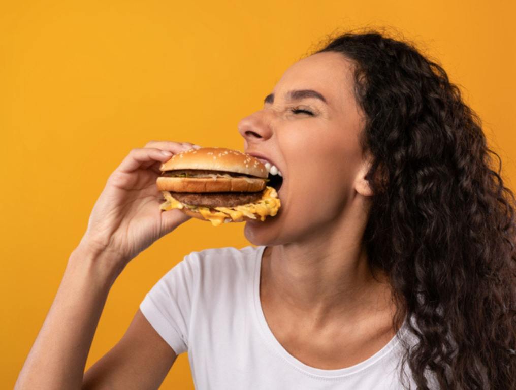 Portrait Of Funny Hungry Lady Biting Burger Eating Junk Food Posing Over Yellow Orange Studio Background.