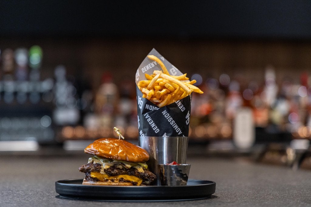 A restaurant cheeseburger and fries wrapped in branded paper sit on a skillet in a sports bar.