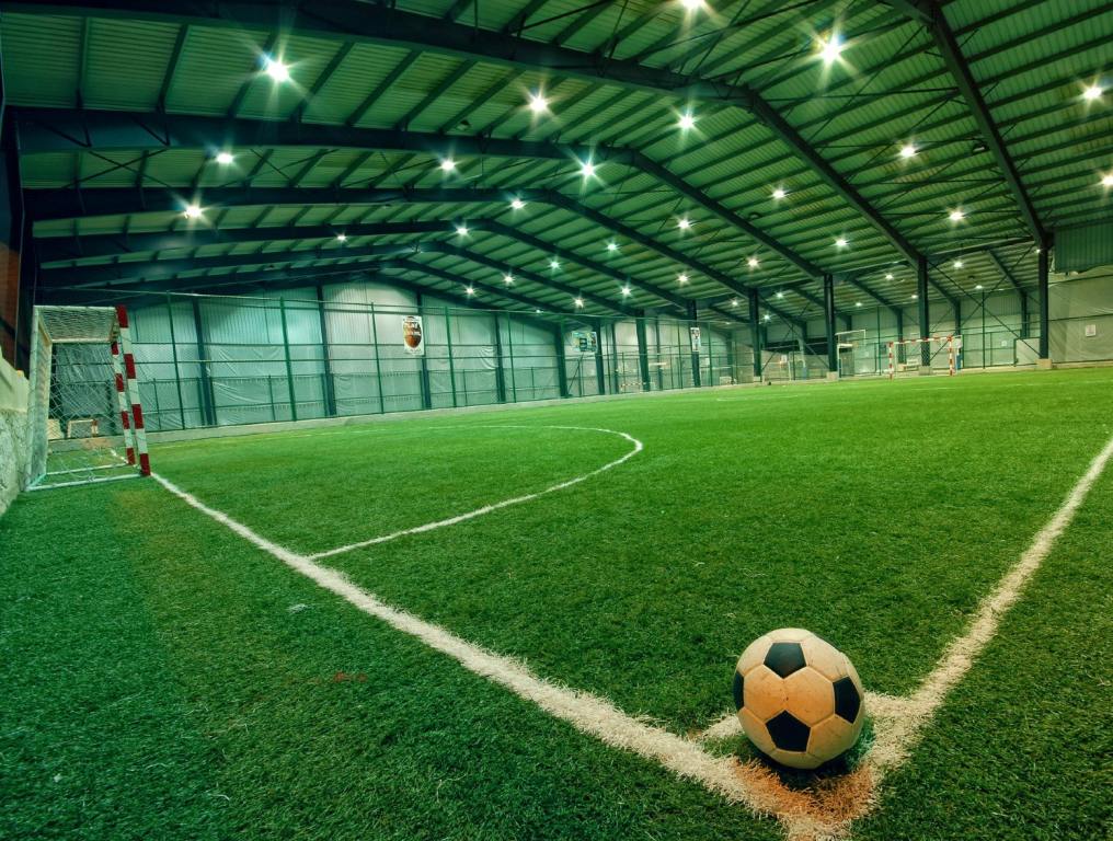 Photo of an indoor soccer field similar to one to be included in the new West Henderson indoor sports facility. The artificial turf is painted green, with white lines painted on and around the field. There is a black and white soccer ball placed in the corner kick space closest to the camera.