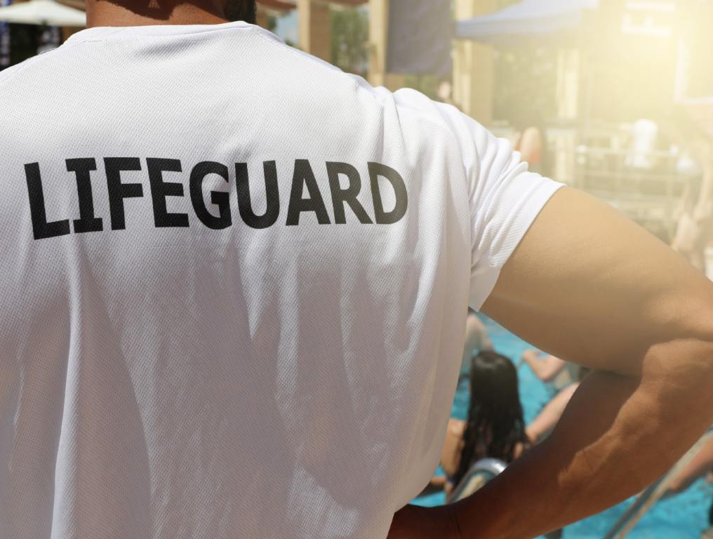 The City of Las Vegas is hiring lifeguards for municipal pools. Photo of a lifeguard on duty at a public pool. Most of the frame is filled up with this man's back. He is wearing a white t-shirt with the word "LIFEGUARD" in black capital letters printed on the upper back. He is holding his right hand on his right hip while he surveys the bathers in the pool. One girl with long black hair can be seen standing in the pool through the crook in his arm. The upper right corner of the photo is overexposed in the sunlight.