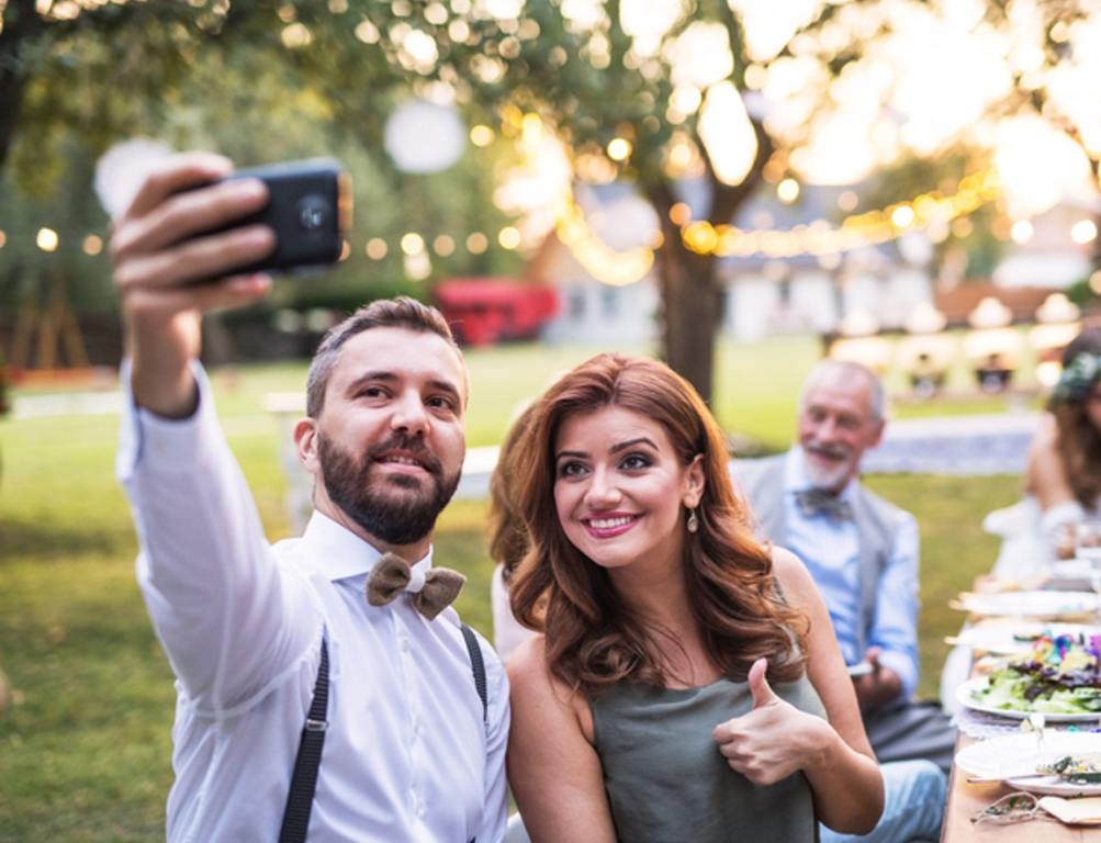 A couple taking selfie at the wedding reception outside in the backyard. Bride, groom and guests sitting at the table.