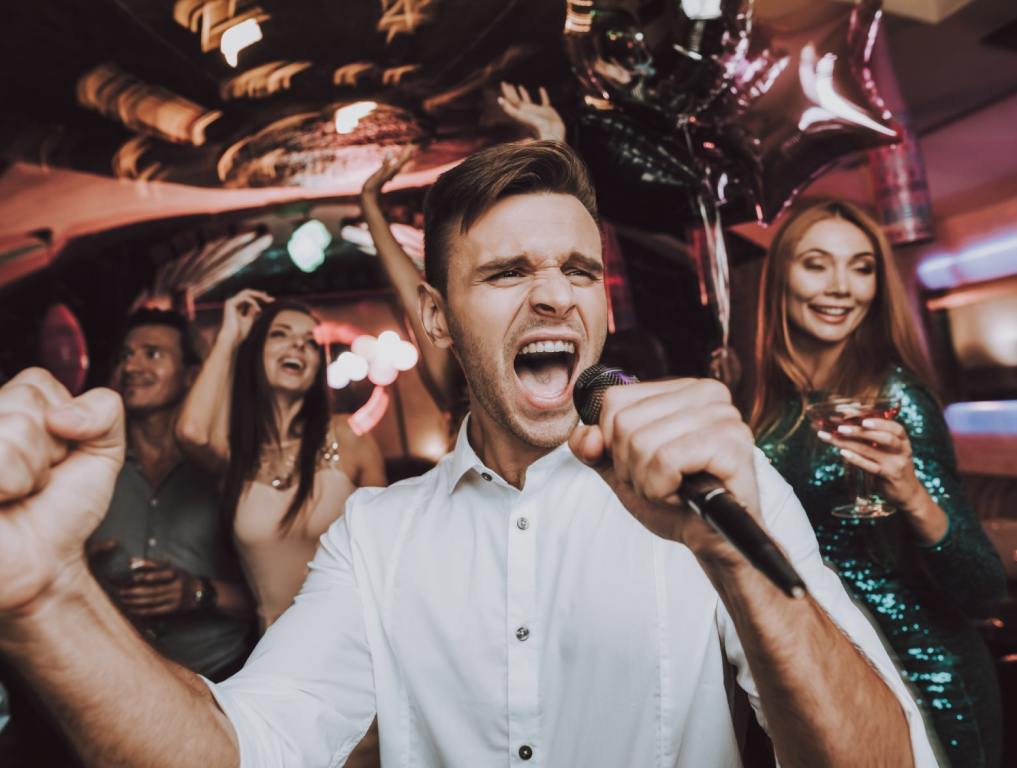 A young white man in a button up shirt is holding a microphone and singing emphatically while his friends look on happily behind him. Concept: Las Vegas Karaoke