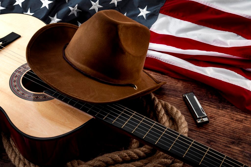 A guitar, cowboy hat, harmonica, and rope lay on a wooden surface next to an American flag.