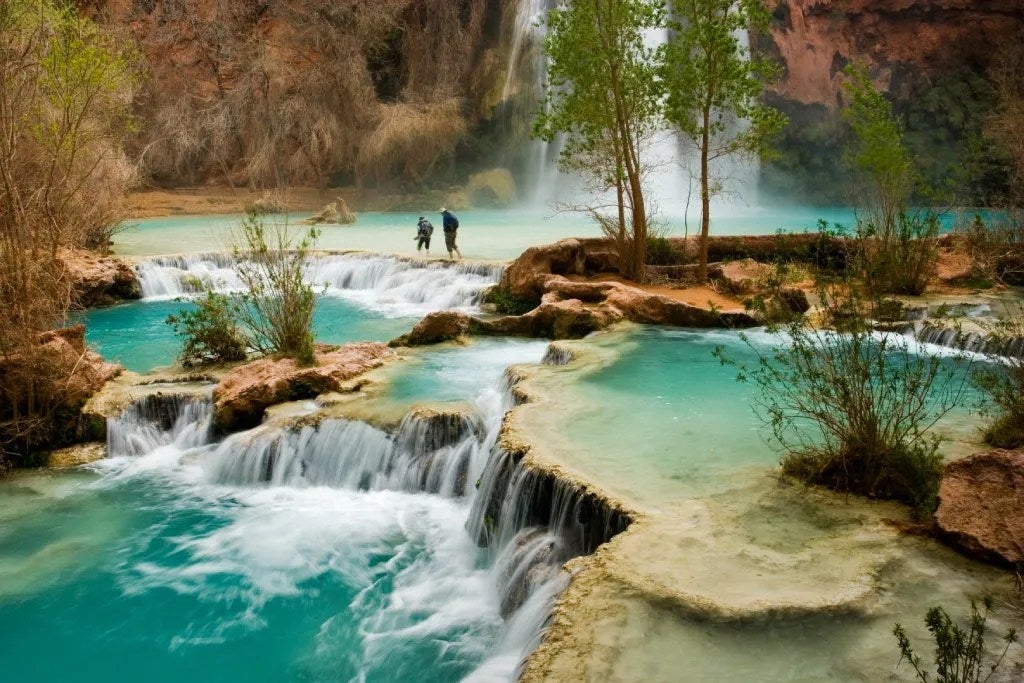 Two people are seen in the distance as they hike through pools of mini waterfalls and a large waterfall in the background.