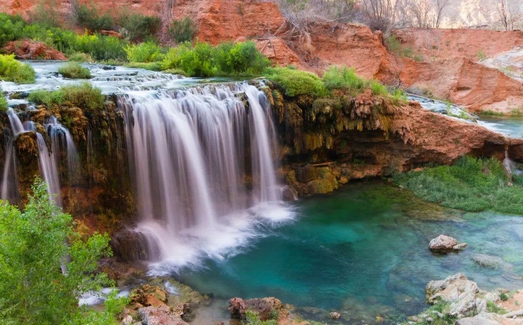 A curtain of water falls over red rocks into a turquoise pool surrounded by trees and rock.