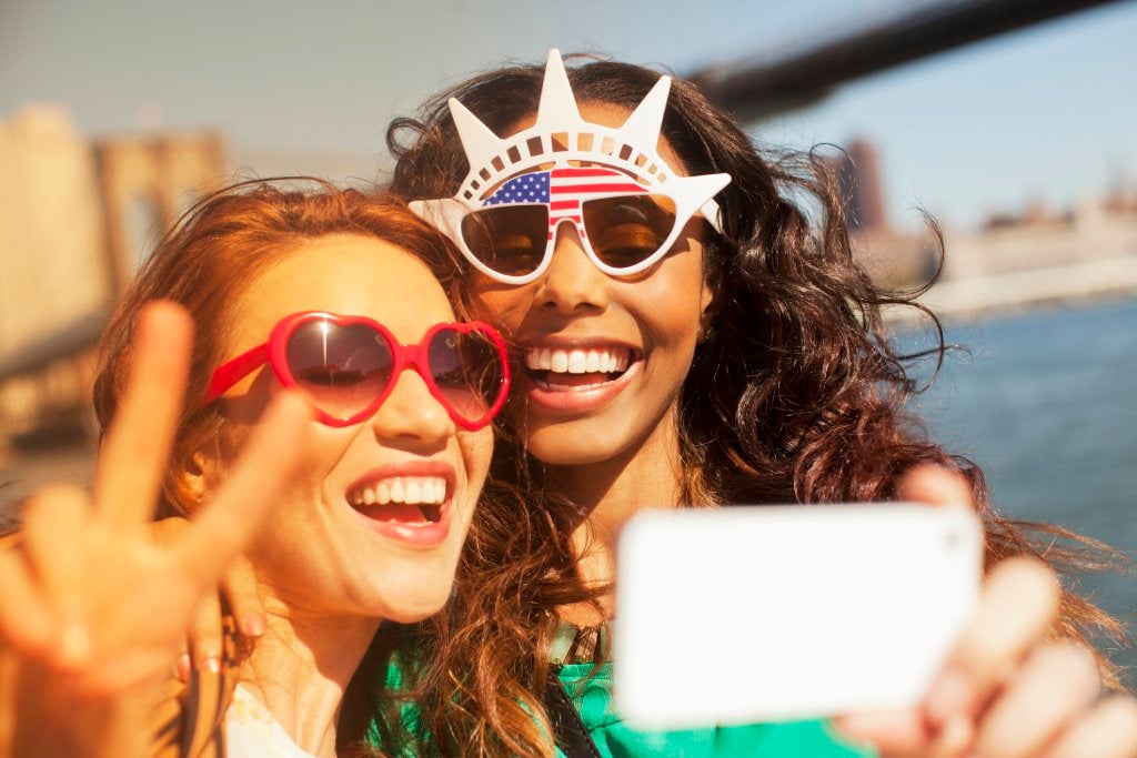 Two women wearing patriotic sunglasses take a selfie with a white cell phone.