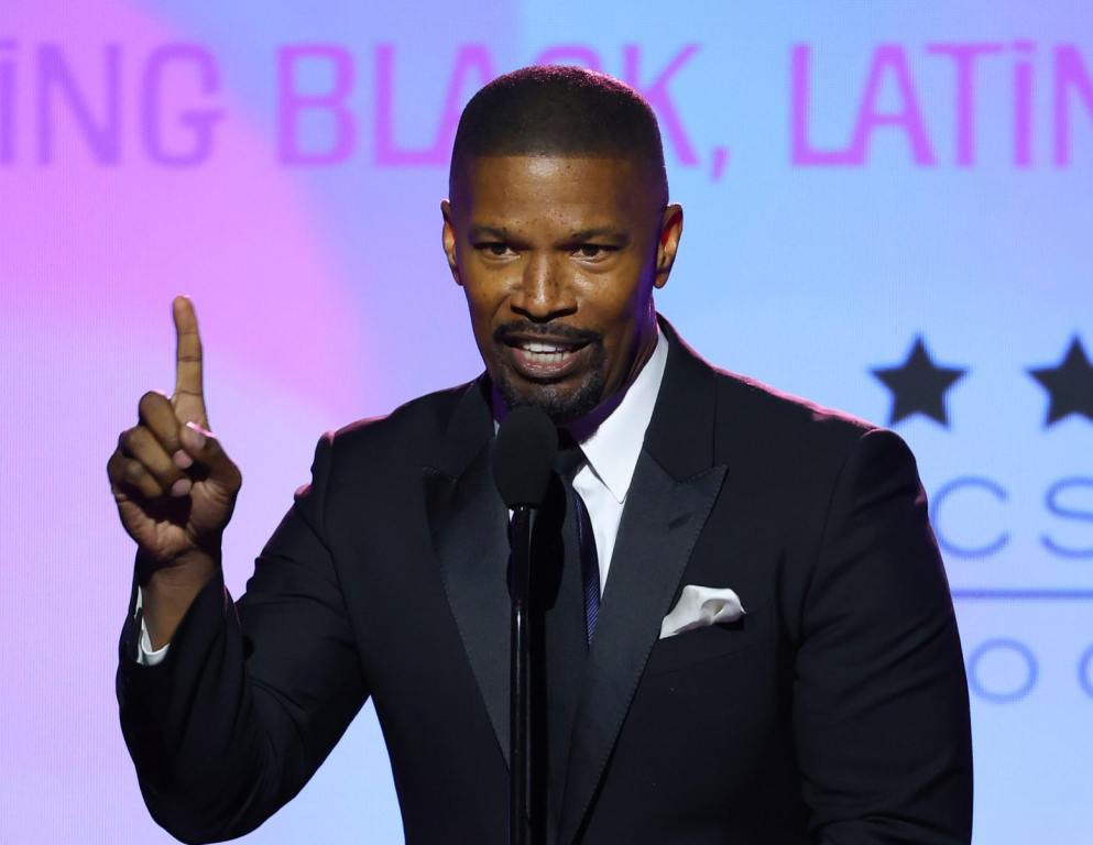 Jamie Foxx accepts the "Vanguard Award" onstage during The Critics Choice Association's Celebration Of Cinema & Television: Honoring Black, Latino And AAPI Achievements pointing a finger up at the podium wearing a black suit and tie, Jamie Foxx Gives Details About Why He Was Hospitalized.