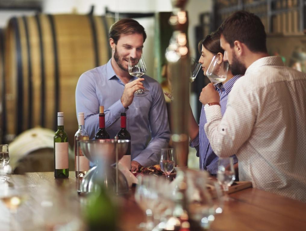 Three white friends at a Las Vegas wine tasting are around a table smelling wine in glasses.