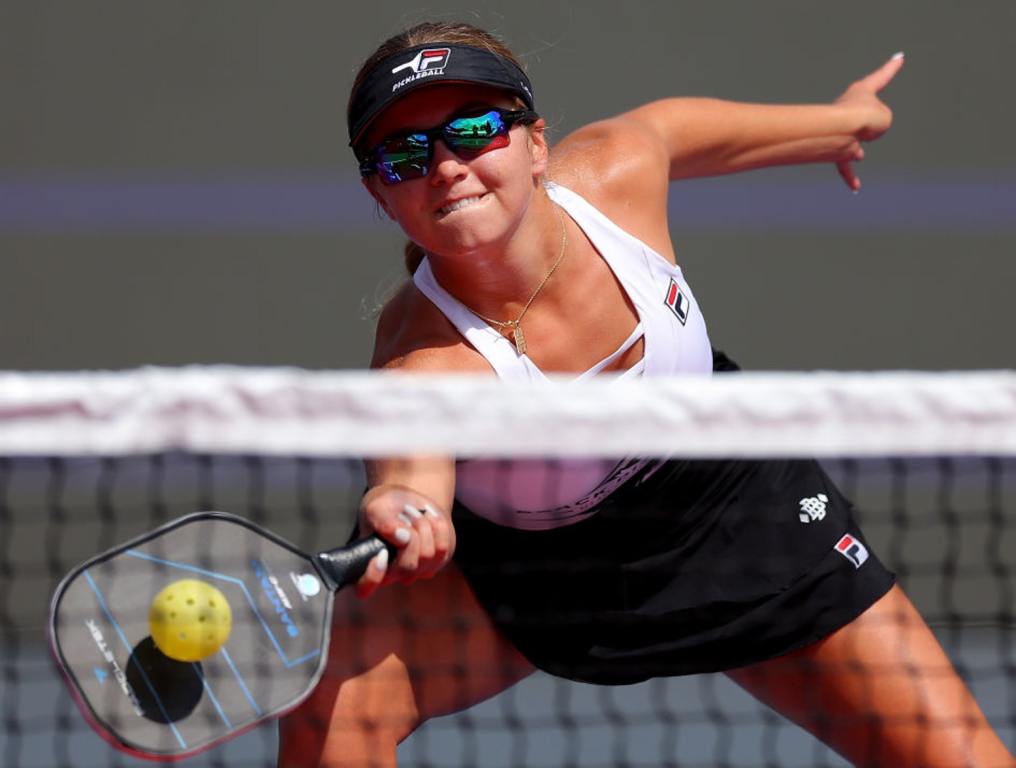 Photo of Anna Leigh Waters, professional pickleball player who will compete in inaugural Las Vegas Pickleball Cup at Fontainebleau Las Vegas August 27 - September 1, 2024. In this photo, she is reaching to her right to complete a return shot of a yellow pickleball. She is wearing a black sun visor, sports sunglasses, a white tank top, a black sport skirt and is reaching to make the shot with her right hand.