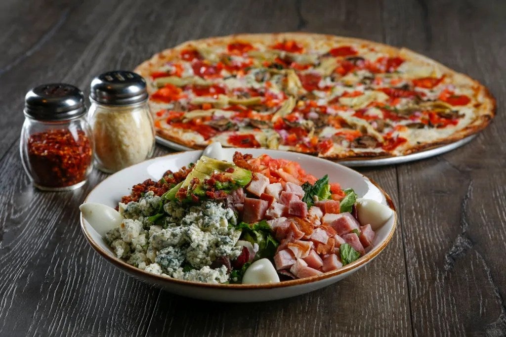 A chopped salad in a shallow white bowl sits in front of a whole pizza on a table. There are parmesan and red pepper shakers to the left.