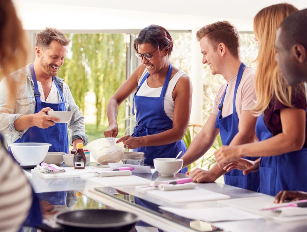 A group of multiracial baking students in blue aprons stand around a kitchen island stirring ingredients in baking bowls. Concept: bread making classes