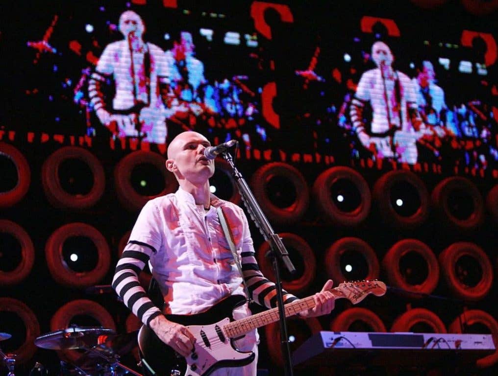 Smashing Pumpkins lead singer Billy Corgan plays guitar and sings on stage wearing a white tee over striped long sleeves.