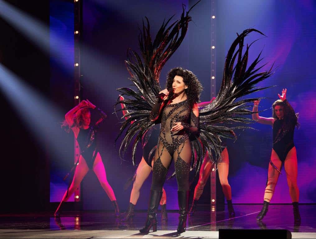 An actress portrays superstar Cher in an iconic black costume with black angel wings. She's on stage singing during The Cher Show.