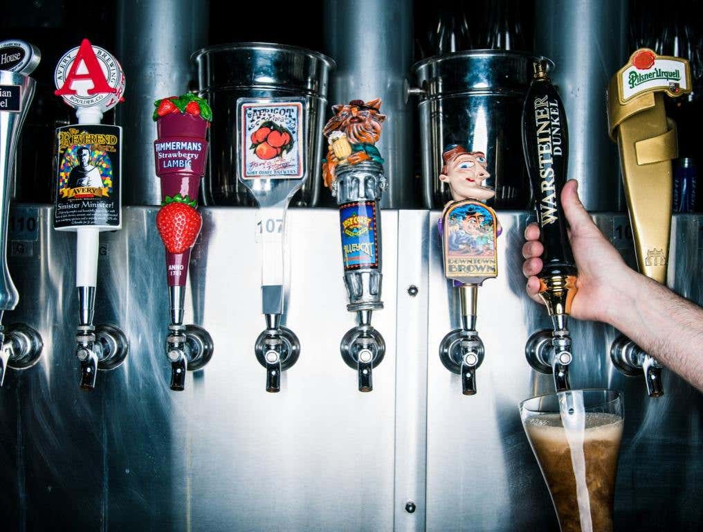 A line of tap beer handles at Yard House restaurant shows a variety of beer labels with bartenders hand shown pouring a beer from one.