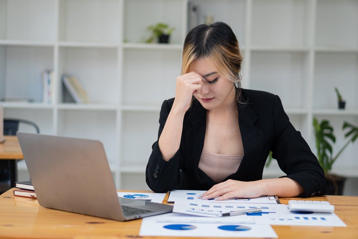 Asian woman with headache working on a laptop and analyzing financial charts on the table