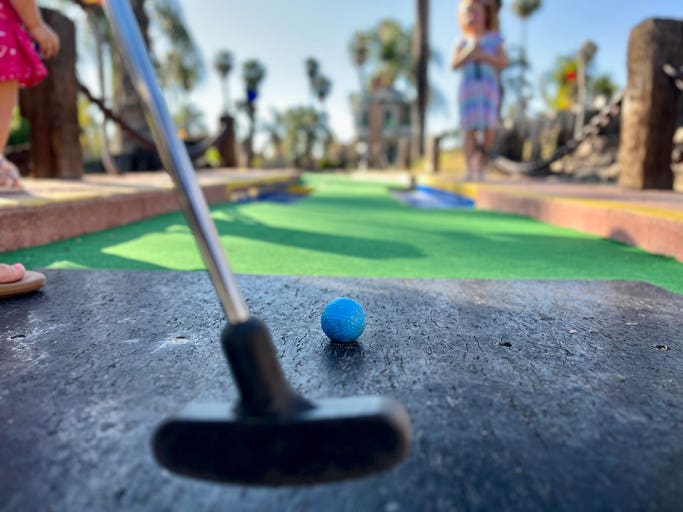 A player on a mini golf course is about to attempt a putt. A family on summer vacation is playing in the evening.