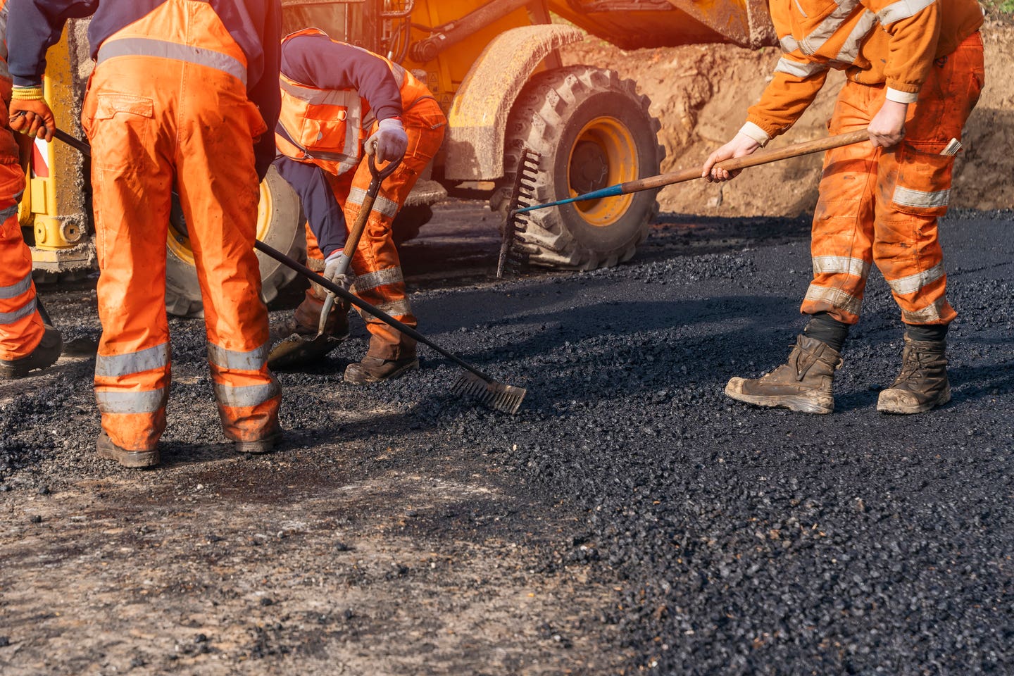 uniforms diligently lay down asphalt on a roadway. Heavy machinery is visible in the background, adding to the busy atmosphere of the worksite.