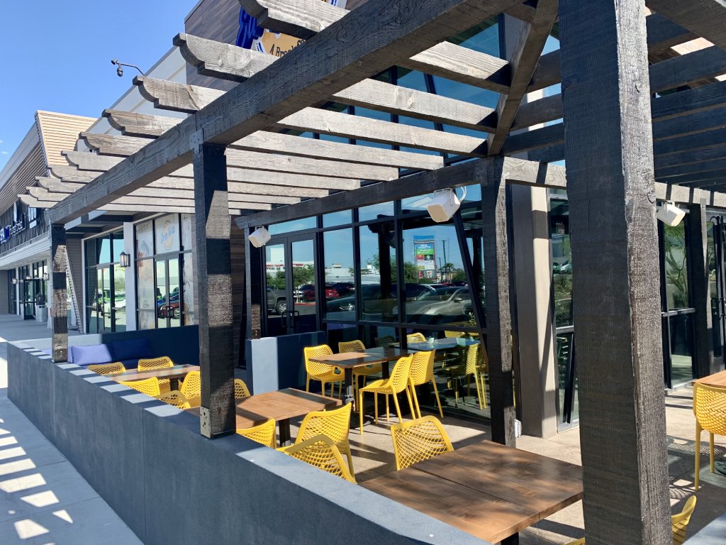 Exterior view of a restaurant patio with yellow dining chairs and open wooden beam ceiling.