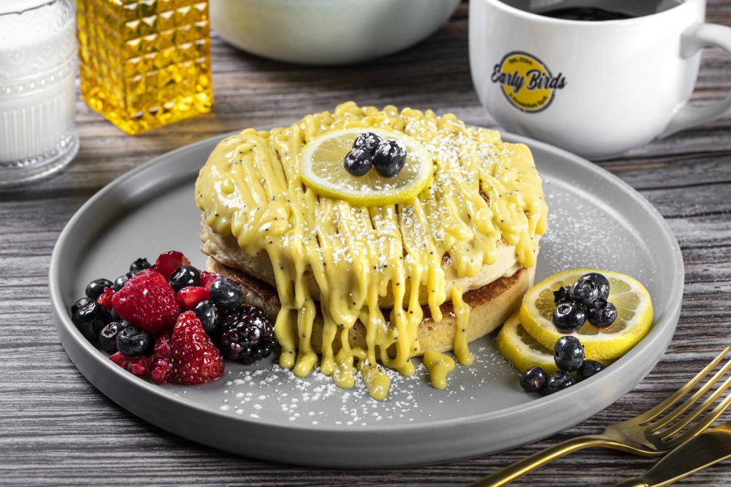A stack of pancakes are covered in a yellow icing drizzle. They are on a plastic gray plate with raspberries and blueberries on the side.
