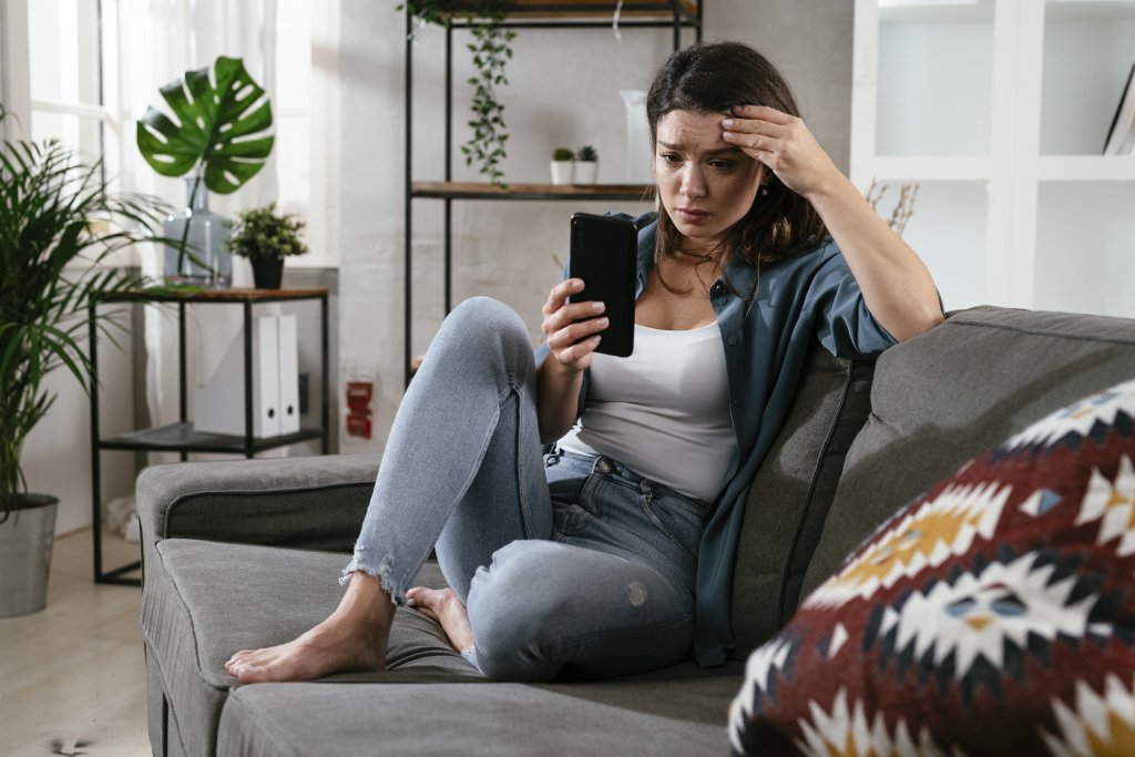 A young brunette woman sitting on a couch is wearing a white tank top, blue button-up and jeans. She is worriedly looking at her phone with her hand on her head.