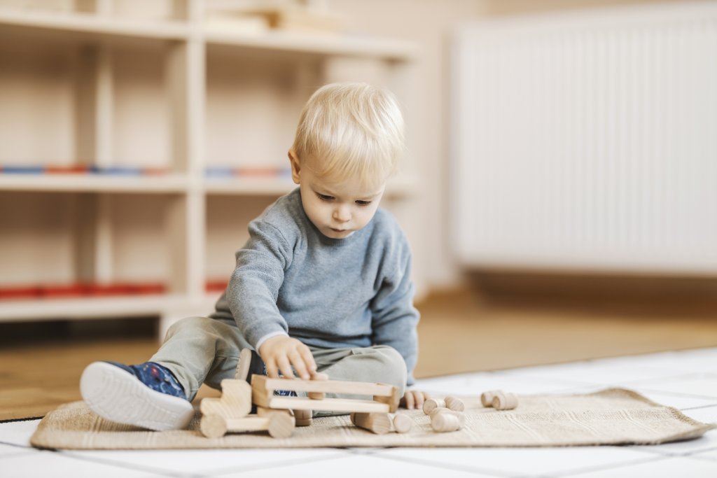 A blonde toddler plays with wooden blocks on the floor of a daycare.