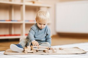 A blonde toddler plays with wooden blocks on the floor of a daycare.